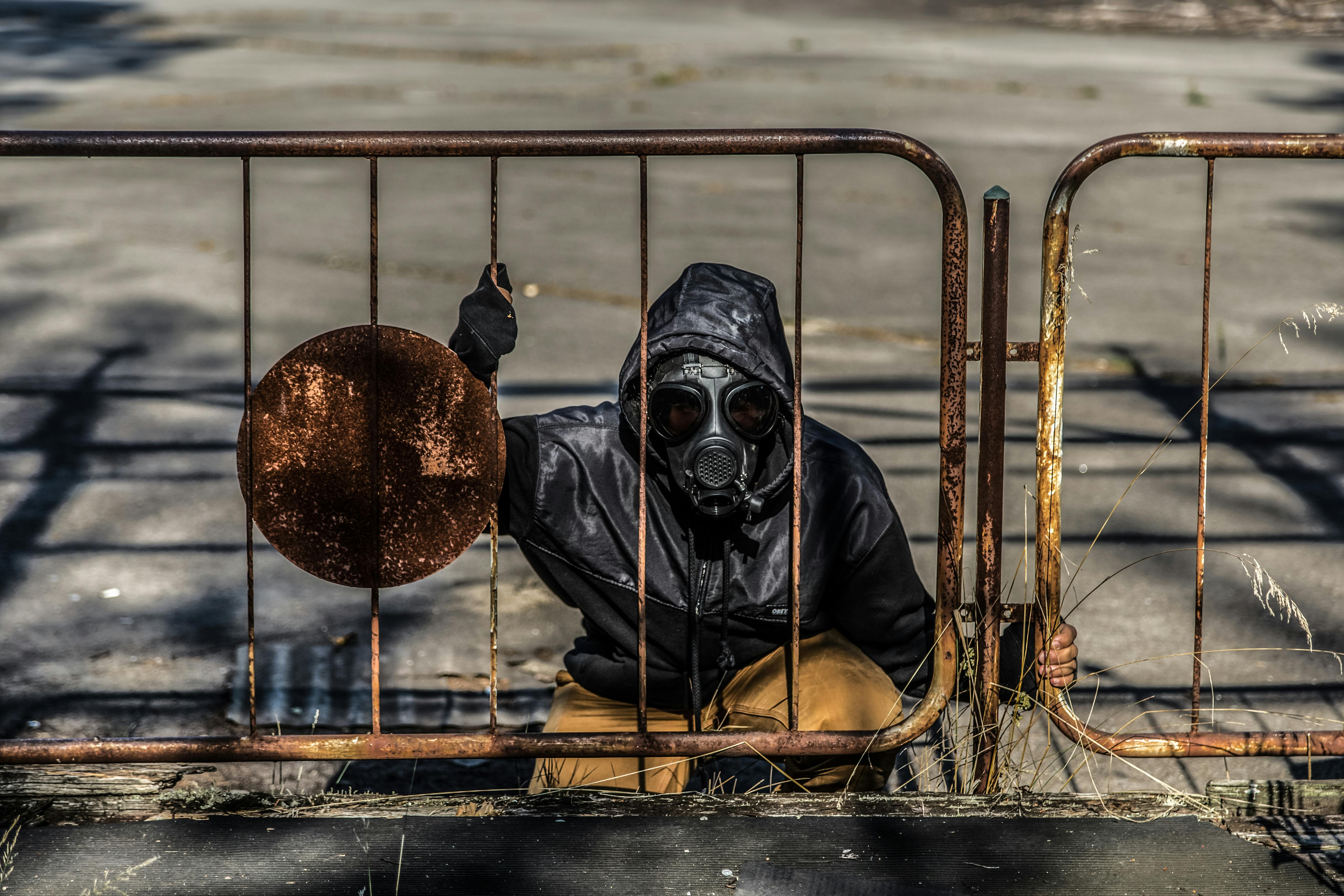 Person wearing a gas mask crouches behind a rusty fence, depicting a post-apocalyptic scene.