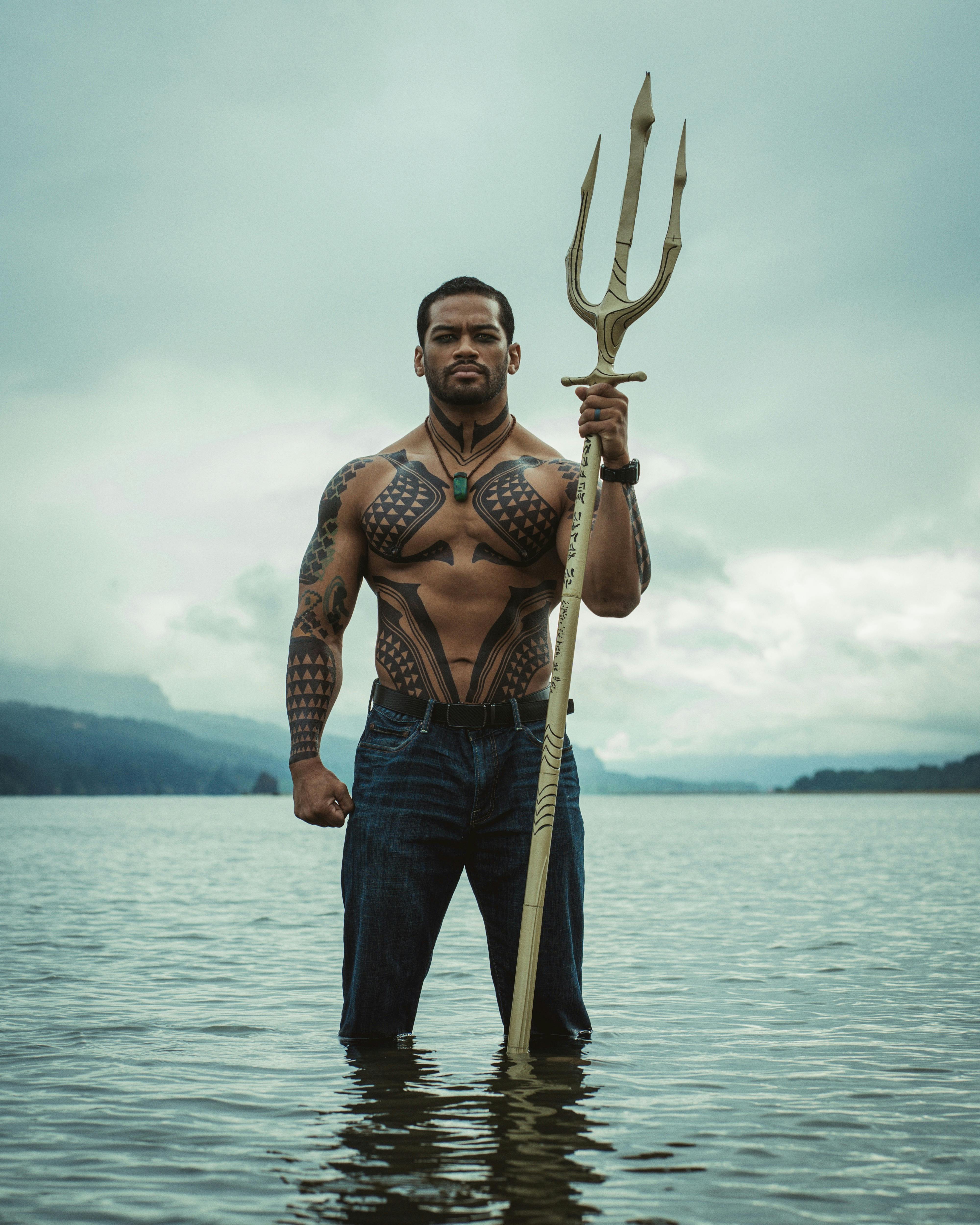 Image of a muscular man with tribal tattoos holding a trident in a serene lake setting.