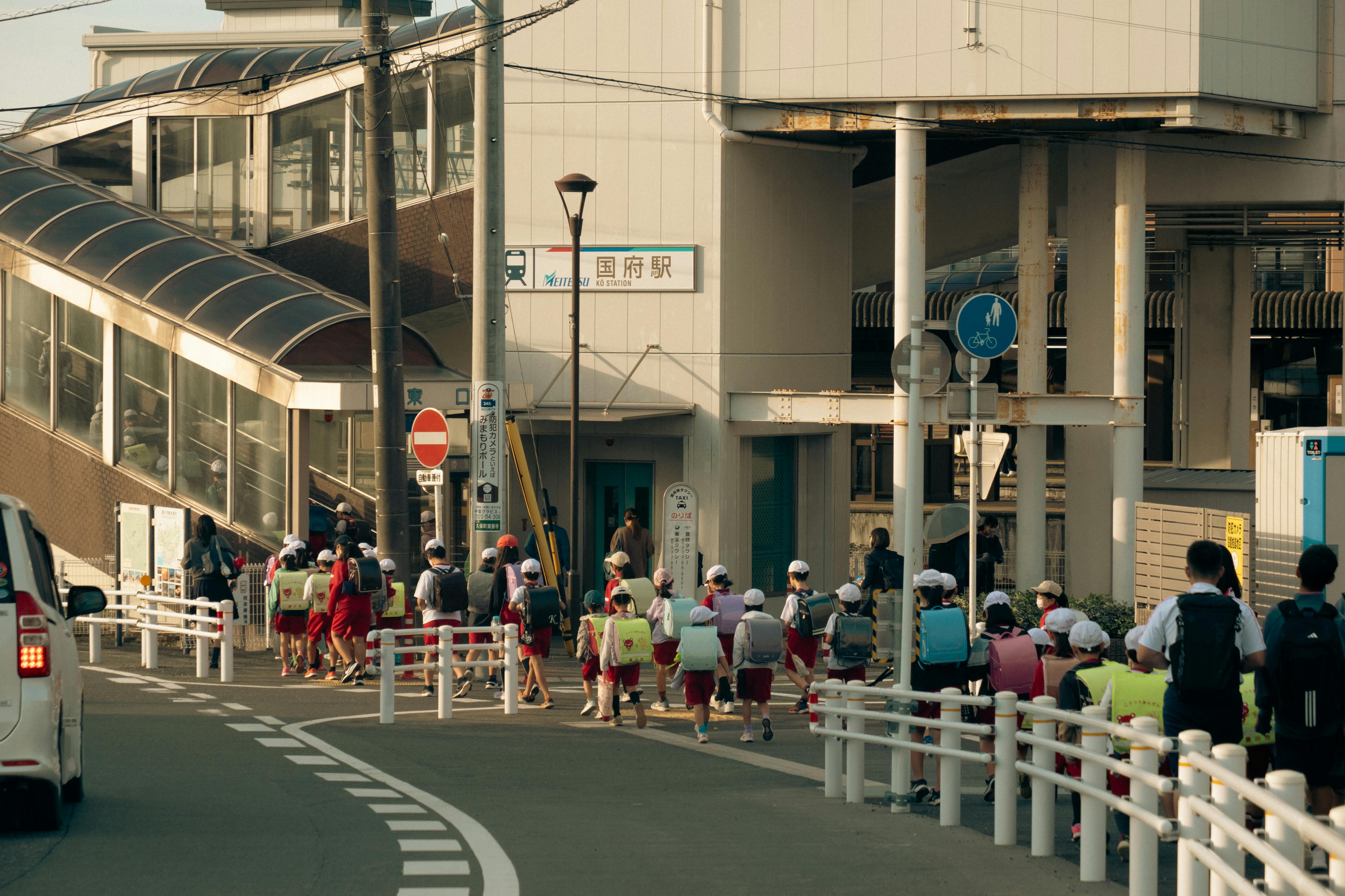 Group of children with backpacks walking towards train station in Toyokawa, Japan, during daytime.