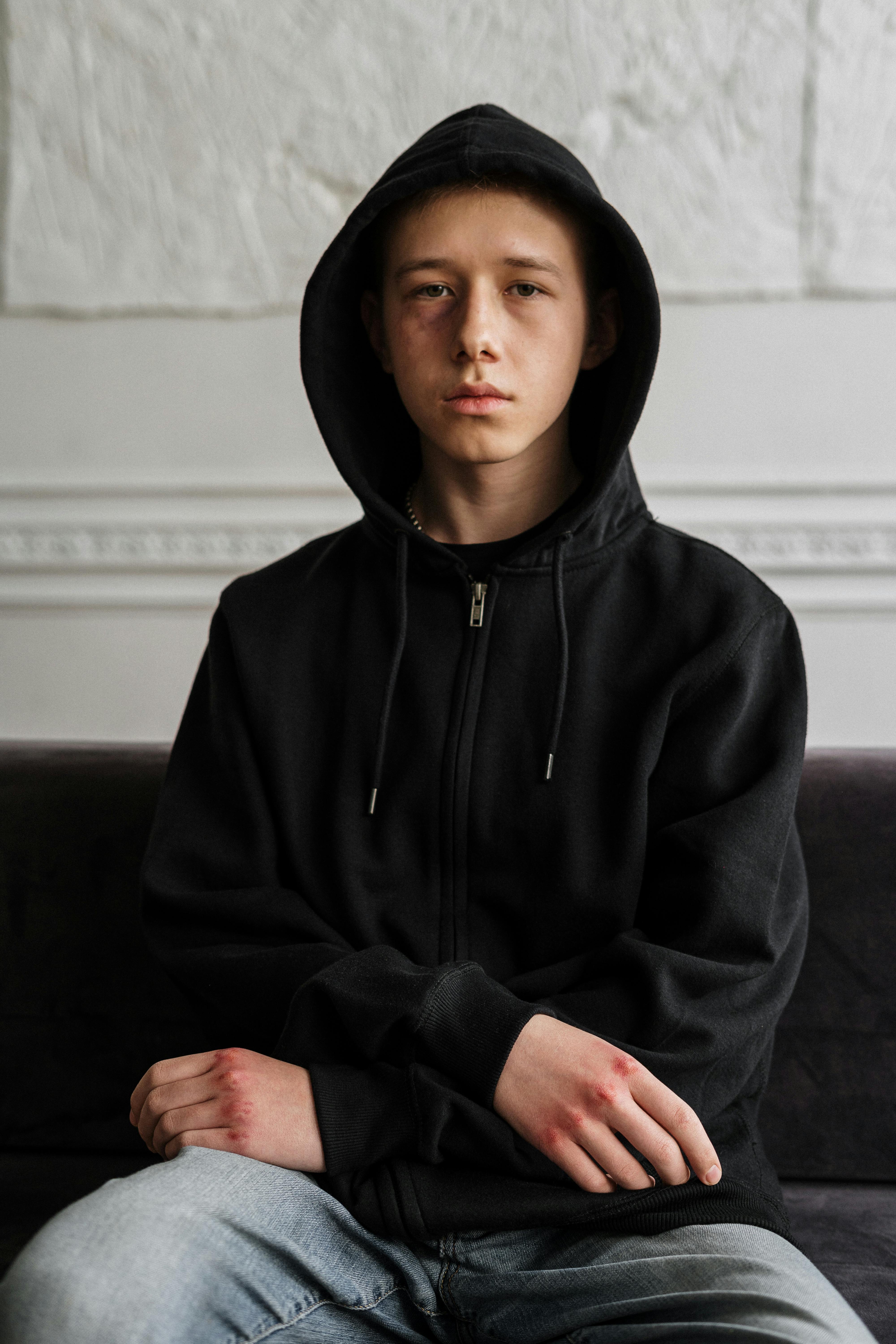 Teenager in black hoodie sitting indoors, contemplating with a serious expression.