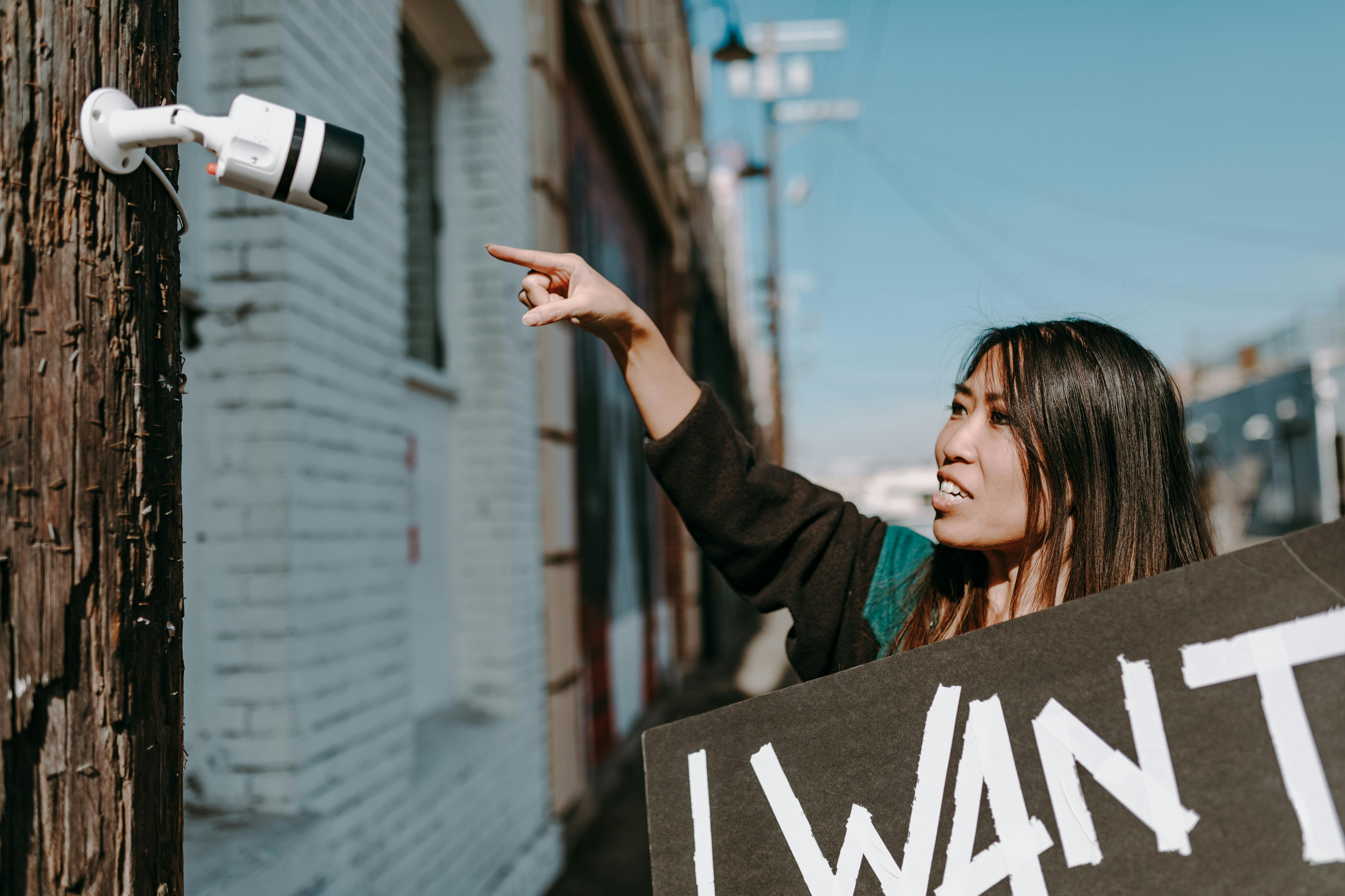 A woman holds a placard, pointing at a CCTV camera during an outdoor protest.